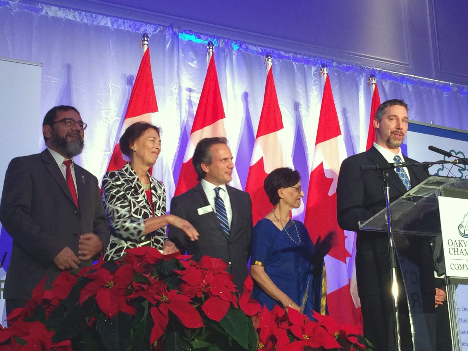 Council leaders on stage beneath the flags while a faith leader speaks at the podium.